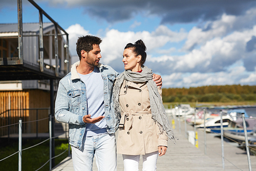 Content handsome young man in jacket walking with beloved woman and embracing her by shoulder, he sharing his story with girlfriend during stroll on pier