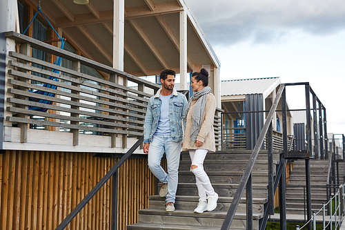 Positive young couple in autumn clothing holding hands and moving down stairs of cottage while chatting outdoors