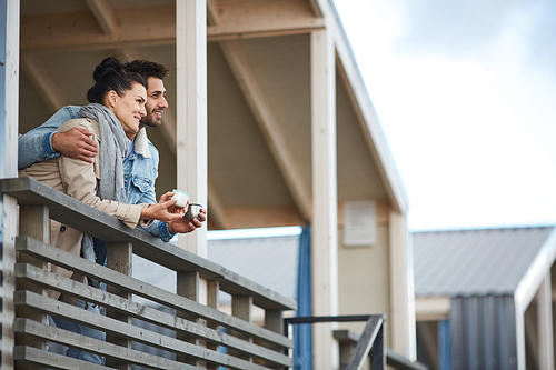 Cheerful dreamy beautiful couple standing on balcony and leaning on railing of cottage while contemplating nature and drinking coffee together