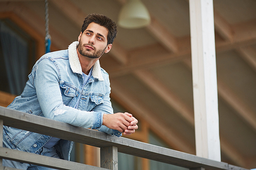 Content dreamy young man in warm denim jacket leaning on railing and looking away while resting on balcony of countryside house