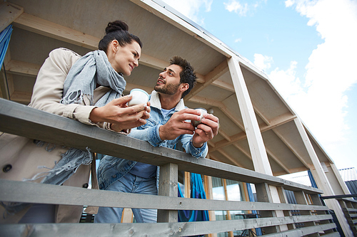 Cheerful smiling beautiful young couple in casual clothing leaning on railing and chatting while drinking coffee on balcony of cottage