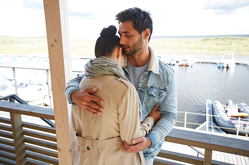 Serious calm loving handsome young bearded man with black hair wearing denim jacket keeping eyes closed and hugging girlfriend at seaport while consoling her