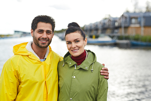 Jolly beautiful young lovers in autumn coats standing on dock and smiling at camera, cheerful bearded man embracing beloved woman