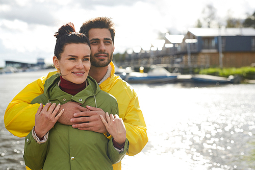 Happy dreamy young couple in coats standing together at seaport and looking at view while contemplating seascape together, smiling man hugging girlfriend