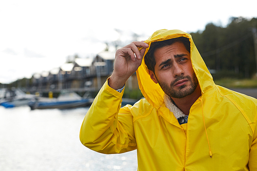 Serious frowning handsome bearded young fisherman adjusting hood of waterproof coat and looking into distance while contemplating seaport on rainy day