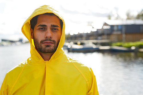Portrait of serious handsome young bearded man in hood wearing waterproof coat standing at dock
