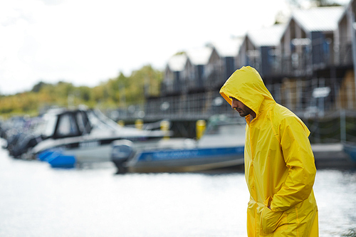 Serious sad young fisherman in yellow waterproof coat holding hands in pockets and looking down while moving along pier at seaport