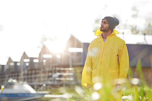 Content pensive young guy in waterproof coat and hat looking around and enjoying sunny day while walking on pier in autumn