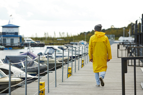 Rear view of lonely young hipster sailor in yellow waterproof coat and hat looking at motor boats and walking over dock in autumn