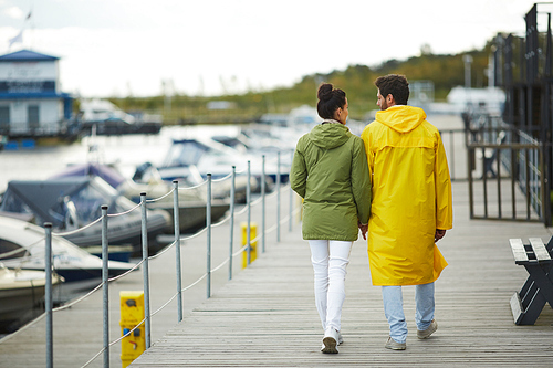 Rear view of young couple in coats holding hands and walking over pier together, they enjoying stroll on autumn day