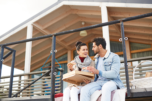 Cheerful excited young couple in casual clothing sitting on blankets and laughing together while eating pizza from box on modern picnic