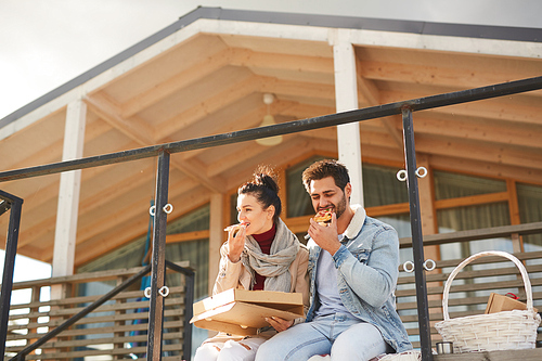 Positive young couple in casual clothing sitting on porch and eating pizza from box on sunny day while resting in country, picnic time