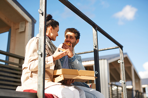 Happy carefree young couple sitting on porch and looking at each other while talking and eating pizza on pier during romantic date