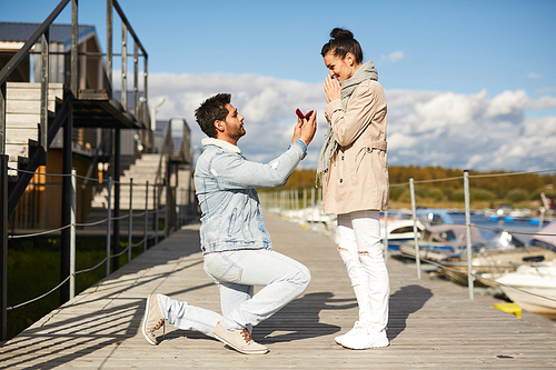 Serious handsome young man in love standing on one knee and holding open jewelry box while proposing to girlfriend on pier