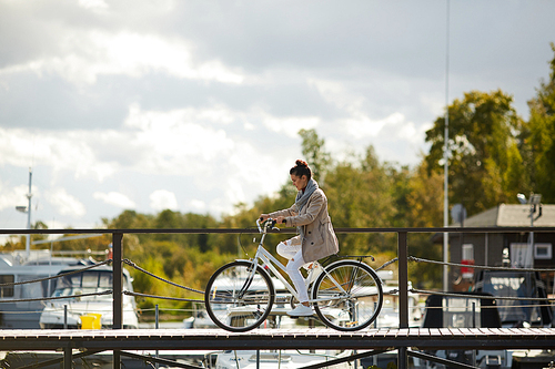 Serious concentrated young woman in coat riding bike carefully on pier and focused on motion while crossing pier alone