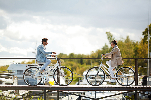 Cheerful young couple riding bikes directly to each other on narrow wooden pier and smiling, they meeting at seaport