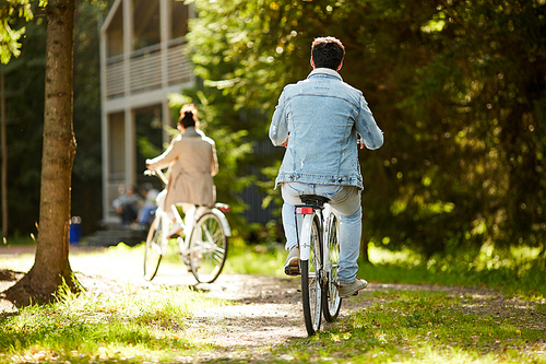 Rear view of playful young couple in casual clothing cycling in forest and racing each other home in warm autumn day