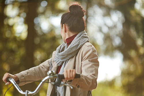 Young woman cyclist in scarf and coat holding handles of bike and turning back while looking around forest in autumn