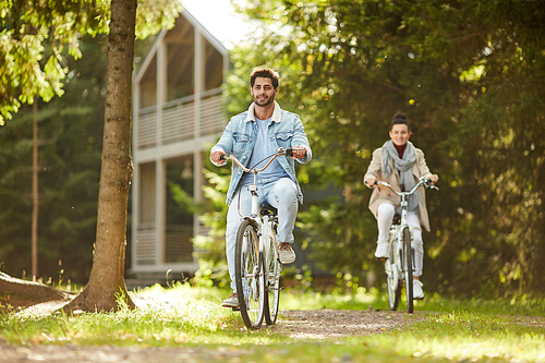 Happy excited young couple in casual clothing enjoying bike ride in forest and having fun during stroll