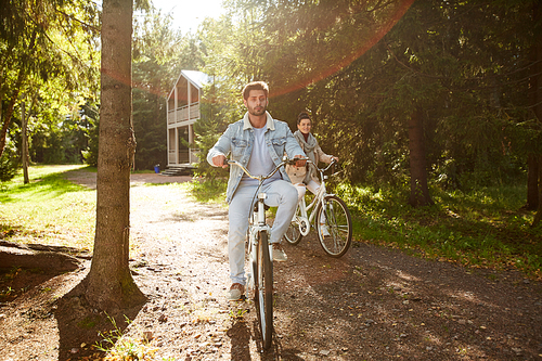 Active couple riding bikes in forest: serious handsome young man in denim jacket riding bicycle on forest path together with cheerful girlfriend