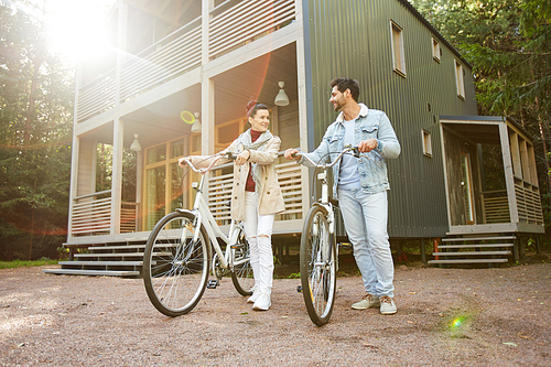 Jolly beautiful young couple in stylish clothing walking bicycles in countryside and looking with love at each other while standing against modern cottage in forest