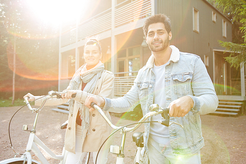 Cheerful beautiful young couple of cyclists in casual clothing walking with bikes together and  in sunlight outdoors