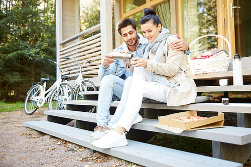 Positive affectionate young couple drinking hot tea from thermos and sitting on step of porch while resting at picnic near house with parked bicycles