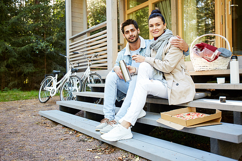 Cheerful beautiful young couple in casual clothing sitting on step with pizza and thermos and smiling at camera, handsome man embracing girlfriend while they having picnic on porch