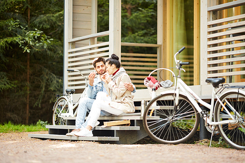 Content young couple sitting on stairs of porch and eating tasty sandwiches while resting after cycling