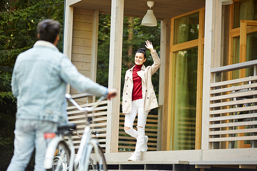 Smiling beautiful young lady in coat standing on porch and waving hand to boyfriend with bicycle while greeting him