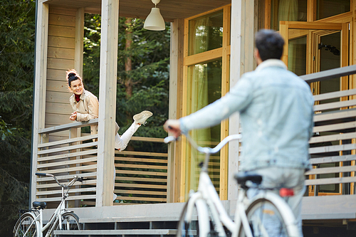 Happy excited young woman in coat leaning on wooden railing and smiling while greeting boyfriend who coming home with bicycle, she waiting for boyfriend in countryside house