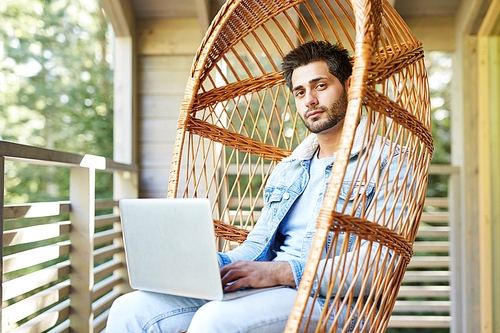 Content confident modern guy with stubble sitting in wicker armchair and  while using pc and typing on keyboard outdoors