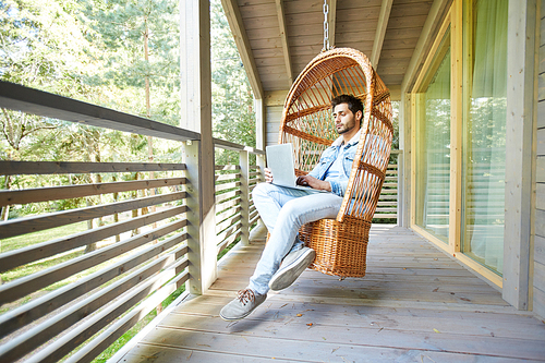 Serious handsome young businessman in casual clothing sitting in hanging armchair and working with laptop on balcony of countryside house