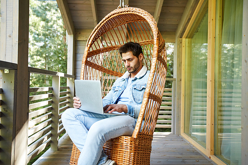 Serious busy young male freelancer in denim jacket sitting in wicker armchair and using laptop while browsing Internet on balcony of cottage house