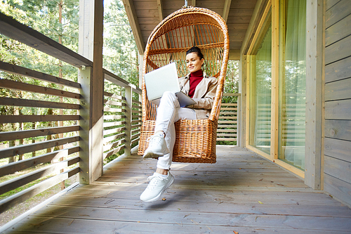 Content confident woman working as freelancer sitting in comfortable hanging chair and using modern laptop on balcony of countryside house