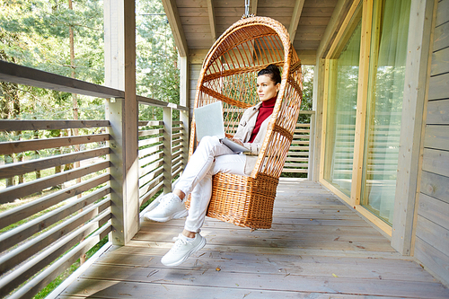 Serious pensive young lady freelancer in white sneakers sitting in hanging wicker chair and using laptop while working on balcony