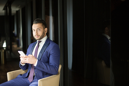 Young elegant businessman in suit sitting in armchair in dark room, texting in smartphone and 