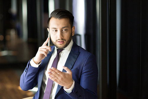 Young confident broker in elegant suit consulting client by smartphone in dark office or room