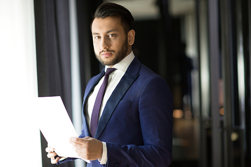 Young elegant businessman with document  while standing by office wndow