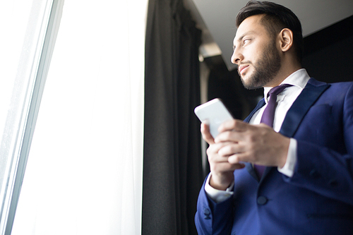 Young pensive manager in suit looking through office window while texting in smartphone