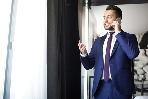 Young and confident broker in formalwear standing by office window, looking through it and talking to client or partner by smartphone
