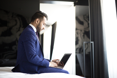 Young businessman sitting in hotel room with laptop in front and searching for online information about new companies