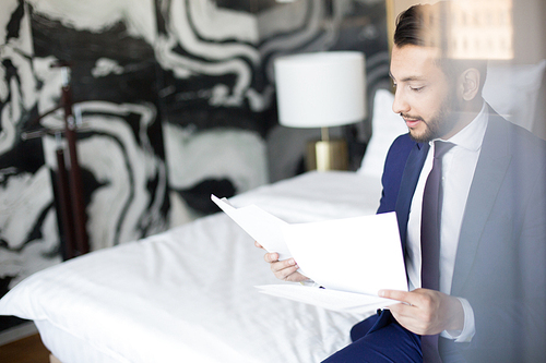 Young business traveler sitting on bed in hotel room, reading financial documents and preparing for meeting with partner