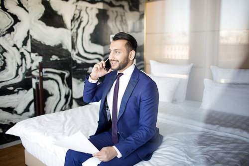 Young cheerful economist in formalwear sitting on bed in hotel room and talking to clients on smartphone