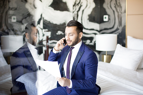 Successful broker or financier in suit reading terms of new financial contract to business partner by smartphone while sitting on bed in hotel room