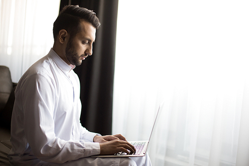 Young businessman in white shirt sitting by window in hotel room and browsing in the net