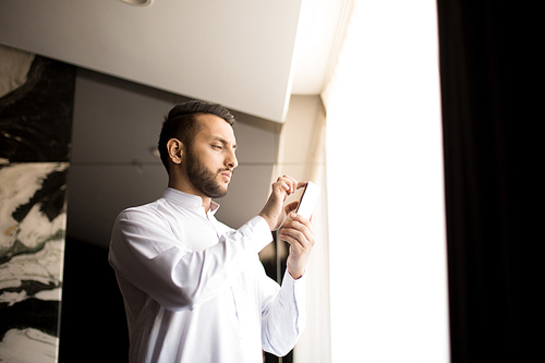 Serious guy volumizing image in smartphone while standing in hotel room by window