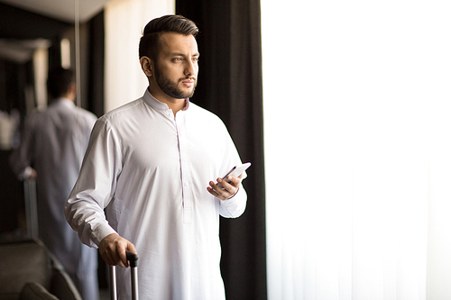 Bearded guy in white looking through window while going to leave hotel room for airport