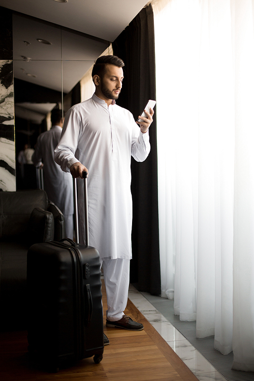 Contemporary young man or businessman with smartphone and baggage standing by window in hotel room