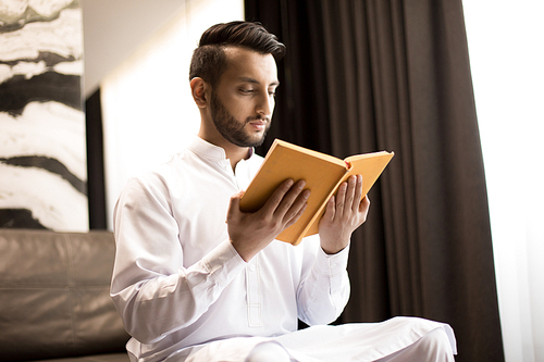 Young handsome man in white clothes reading book in yellow cover while sitting in the room
